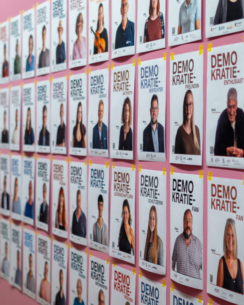 Viele einzelne Plakate hängen an einer rosa Wand mit der Aufschrift "Demokratie Vertreter" im mpk Museum Pfalzgalerie Kaiserslautern