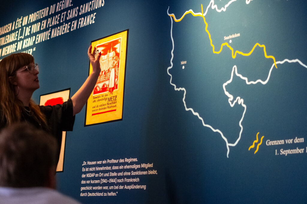 Ausstellungsansicht im mpk Museum Pfalzgalerie Kaiserslautern der Ausstellung "Zeitsprung: Gekauft. Getauscht. Geraubt?". Kuratorin und Provenienzforscherin Laura Vollmers mit Brille, dunkler Kleidung und langen, rötlich-blonden Haaren steht vor einer blau gestrichenen Wand und erklärt mit erhobener Hand ein historisches Propagandaplakat aus der Zeit des Nationalsozialismus. Das Plakat zeigt eine stilisierte Ansicht von Metz in roten und gelben Farbtönen mit dem Text: „Besuchen Sie das alte, ehrwürdige Metz und die schönen Moseltäler unserer deutschen Westmark“. Rechts auf der Wand ist eine Karte mit gelb und weiß eingezeichneten Grenzen und Städten wie „Metz“ und „Saarbrücken“ zu sehen. Darunter befinden sich französisch- und deutschsprachige Zitate über NS-Aufarbeitung.
