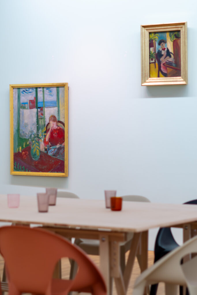 Ausstellungsansicht mpk mit Gemälden an der Wand vor denen ein Tisch mit bunten Gläsern steht.