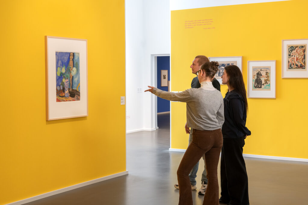 Besucher:innen betrachten fasziniert ein expressionistisches Gemälde in der leuchtend gelben Ausstellungshalle von „Unerhört Expressiv! Lebensgefühle von Kirchner, Kollwitz und Co“ im mpk Museum Pfalzgalerie Kaiserslautern – ein inspirierendes Kulturerlebnis für Kunstliebhaber:innen.