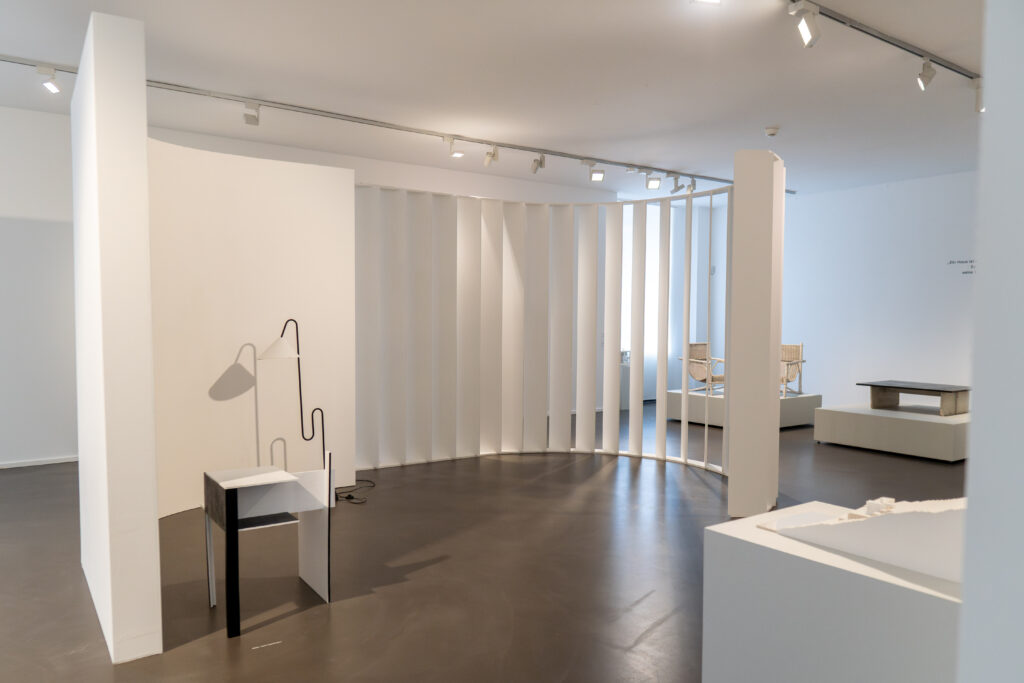 Geschwungene weiße Lamellenwand mit moderner Leuchte und minimalistischen Möbelobjekten in der Ausstellung „Golden Girls No. 3: Eileen Gray“ im mpk Museum Pfalzgalerie Kaiserslautern mit Fokus auf modernes Design und Raumgestaltung.
