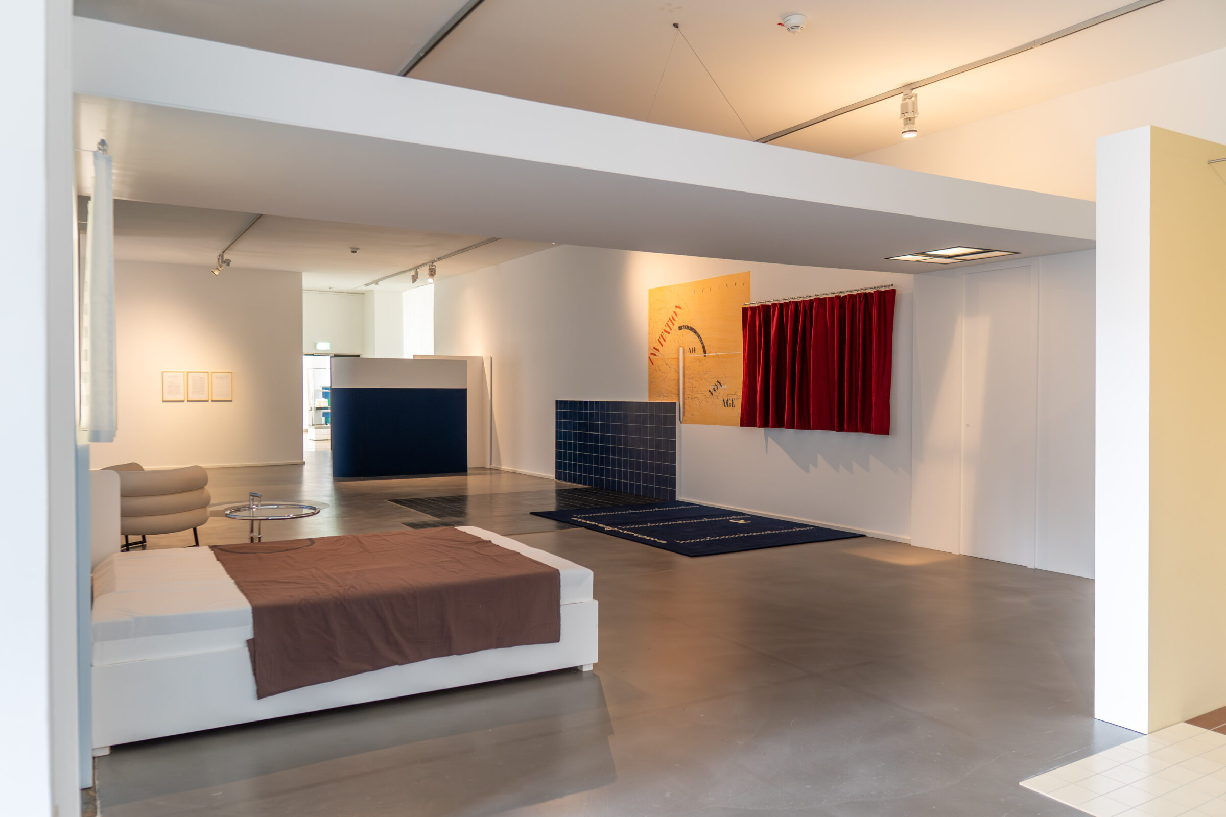 Modernistische Rauminstallation mit Bett, Teppichen, roten Vorhängen und dunkelblauen Fliesen in der Eileen-Gray-Ausstellung im mpk Museum Pfalzgalerie Kaiserslautern als