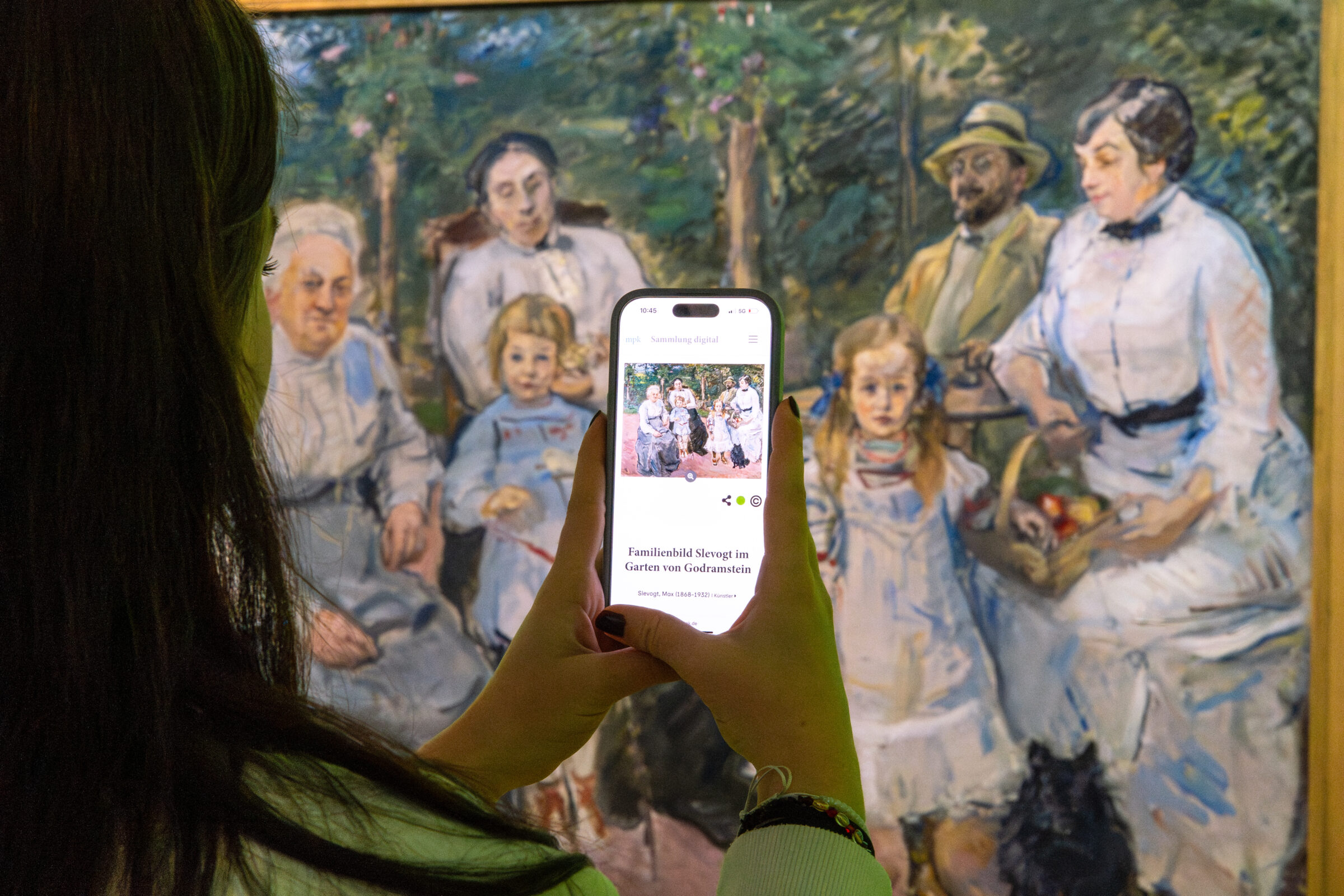 Besucherin nutzt ein Smartphone vor einem Gemälde im mpk Museum Pfalzgalerie, um über die digitale Sammlung Zusatzinformationen zu einem historischen Max Slevogt Familienporträt aus der Museumssammlung abzurufen.