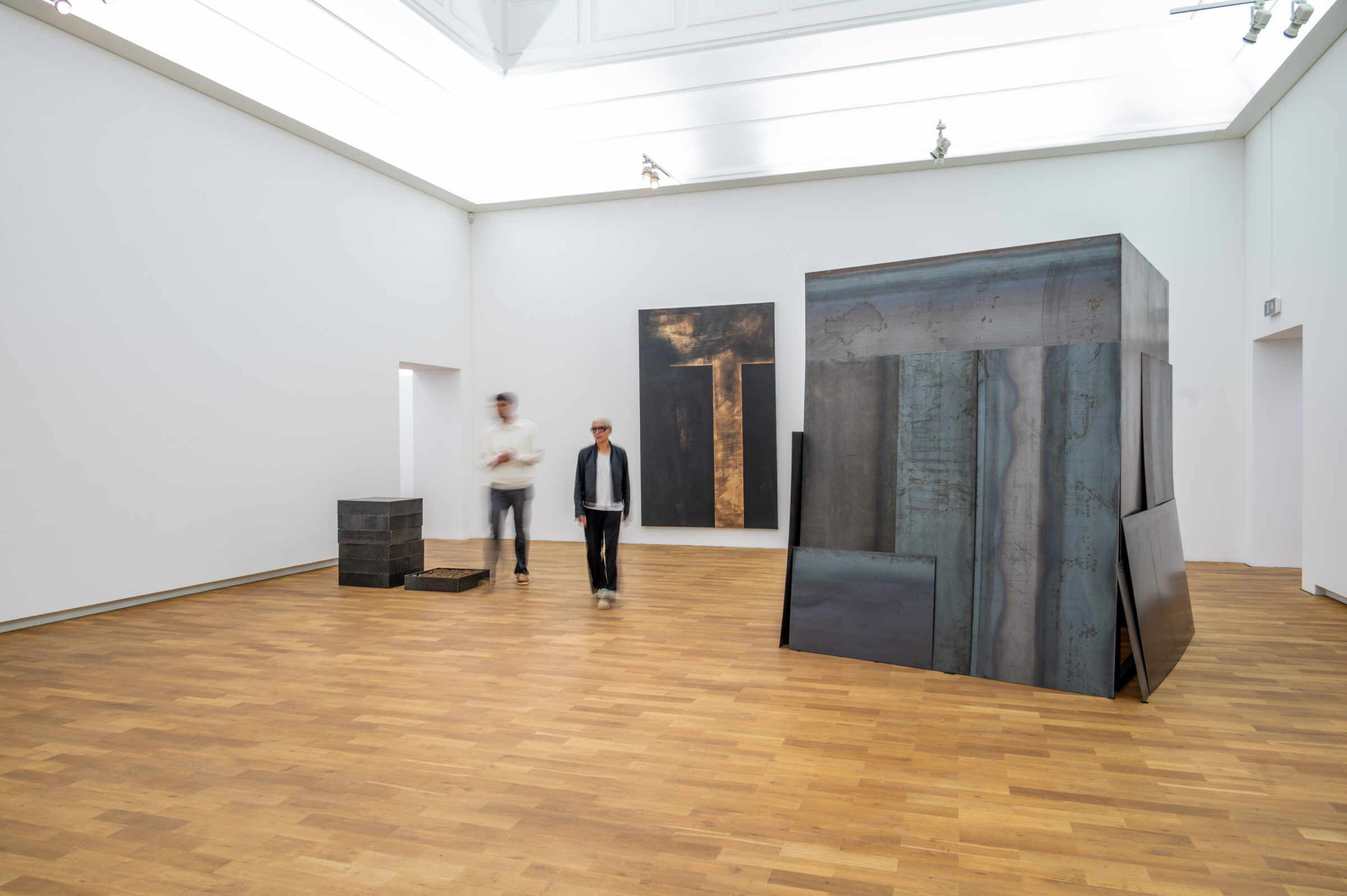 Blick in die Ausstellung „Madeleine Dietz: Erde, Stahl und Licht“ im mpk Museum Pfalzgalerie Kaiserslautern. Der großzügige, lichtdurchflutete Museumsraum mit weißen Wänden und hellem Parkettboden zeigt mehrere großformatige, abstrakte Kunstwerke der Künstlerin Madeleine Dietz. Rechts steht eine monumentale, begehbar wirkende Konstruktion aus grauen Stahlplatten. Im Hintergrund hängt ein dunkles Wandwerk mit goldfarbenen Akzenten. Zwei Personen bewegen sich durch den Ausstellungsraum. Die zeitgenössische Kunst von Madeleine Dietz verbindet geometrische Formen mit Materialien wie Stahl, Erde und Licht.